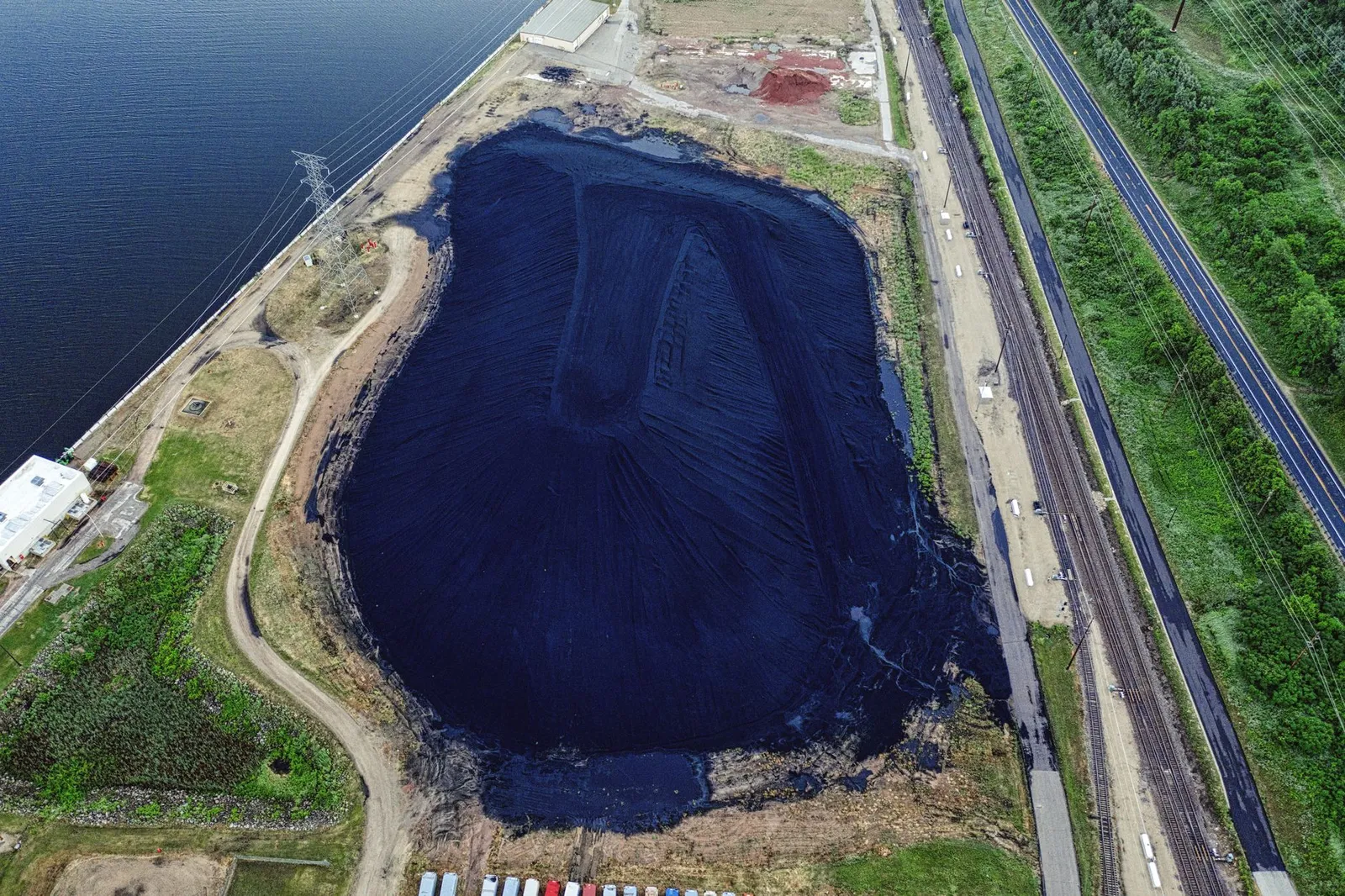 Aerial view of a coal ash pond beside a roadway and rail lines, with a nearby industrial facility