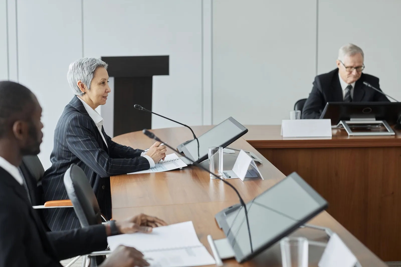 Officials seated at a hearing table with microphones and screens while a speaker addresses them